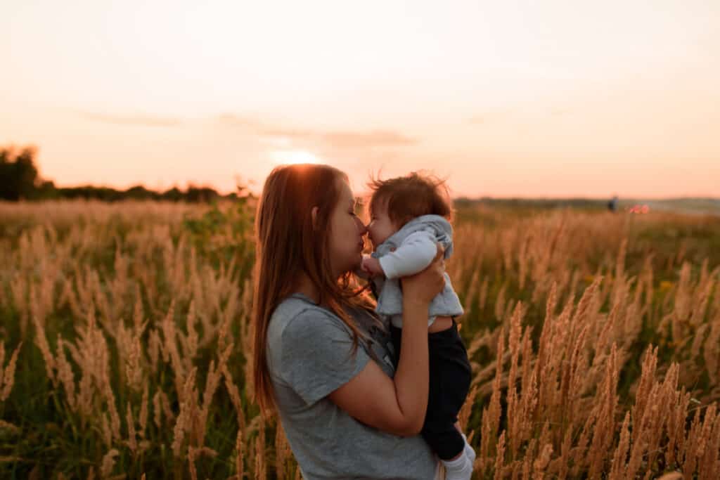 mom snuggling baby sun in field with sunset in background